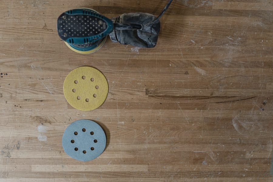 Yellow and blue hook-and-loop sanding discs laid out on a worn wooden bench beside a random-orbit sander